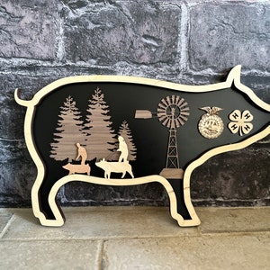 Op de afbeelding: Een houten uitsnijding van een varken met een zwarte achtergrond. Binnenin het varken bevinden zich uitsnijdingen van bomen, mensen, varkens, een windmolen en twee badges. Een badge zegt "FFA" en de andere heeft een klavertje vier.