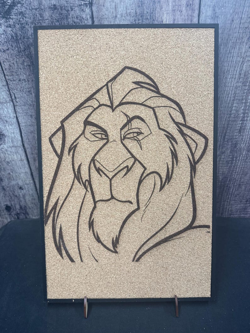 Puede incluir: Un corcho enmarcado con un dibujo lineal negro de la cabeza de un le&oacute;n. La cara del le&oacute;n est&aacute; detallada con una melena, ojos y nariz. El marco es negro y el corcho es de color marr&oacute;n claro.