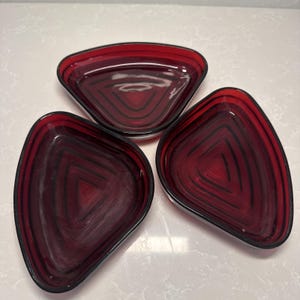 May include: Three triangular, ruby red glass dishes with black linear designs. The dishes have a glossy finish and a slightly irregular shape. The design creates a spiral effect within each dish. The dishes are arranged on a white surface.