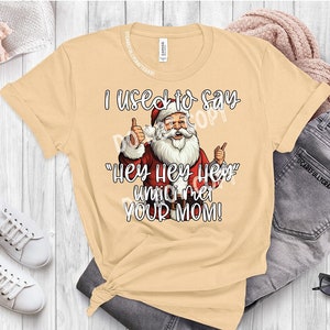 Puede incluir: Una camiseta beige con un gráfico de Papá Noel y el texto "Solía decir "Hey Hey Hey" hasta que conocí a tu madre!"