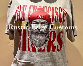 May include: Gray t-shirt with a red and white graphic of a bearded man wearing a red beanie with the San Francisco 49ers logo. The text "SAN FRANCISCO" and "49ERS" are in red. The shirt also has the text "Rustic Edge Customs".