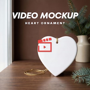 May include: A smooth white heart-shaped ornament with a gold string loop at the top. The text VIDEO MOCKUP HEART ORNAMENT is displayed above the ornament, and a red clapperboard icon is positioned over the centre of the white heart surface.