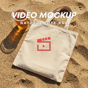 May include: A natural canvas tote bag with the text "VIDEO MOCKUP" and "NATURAL TOTE BAG" in white. The bag has a red graphic of a film clapper with a play button. A clear plastic cup with a light brown liquid sits next to the bag.