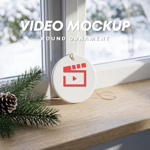 May include: A white round ceramic ornament featuring a red film clapperboard icon in the center. The ornament hangs by a thin gold string. The text VIDEO MOCKUP ROUND ORNAMENT is displayed in white capital letters above the ornament.