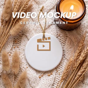 May include: White ceramic ornament with a gold-colored video icon and the text "VIDEO MOCKUP CERAMIC ORNAMENT". The ornament hangs from a gold string. The background includes wheat stalks, a lit candle, and a cream-colored knitted blanket.