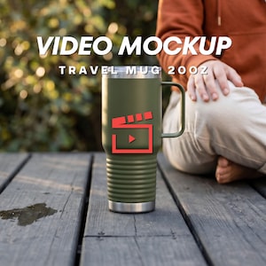 Op de afbeelding: Een olijfgroene reismok met een handvat, voorzien van een rood filmklapper-icoon op de voorzijde. De tekst VIDEO MOCKUP TRAVEL MUG 20OZ staat boven de mok. De mok heeft een geribbeld onderste gedeelte en een zilverkleurige basis en rand.
