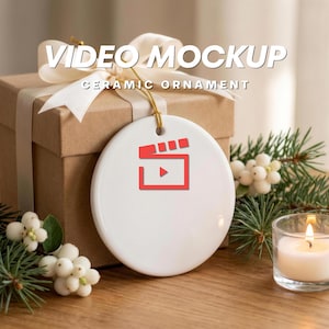 May include: A round white ceramic ornament featuring a red movie clapperboard icon in the center. The ornament is suspended by a gold string against a brown gift box with a white ribbon, accompanied by green pine needles and a small lit candle.