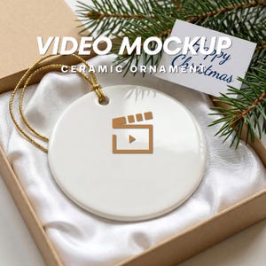 May include: White ceramic ornament with a gold-colored cord, featuring a gold-colored film clapperboard icon. The text "VIDEO MOCKUP CERAMIC ORNAMENT" is printed on the ornament. The ornament is in a gift box with a "Happy Christmas" card.