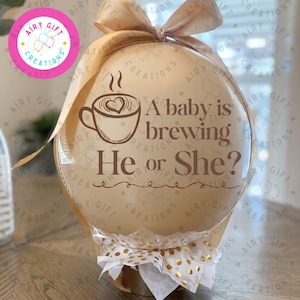 May include: A clear, round balloon filled with a beige balloon, topped with a beige ribbon bow. The surface features a brown graphic of a coffee cup with steam and the text: A baby is brewing He or She? A white and gold polka dot ribbon decorates the base.
