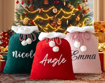 May include: Three large Christmas gift sacks in green, red, and pink, each with a white fluffy trim and a name embroidered in cursive. The sacks have red and white rope ties with white pom-poms. A decorated Christmas tree is in the background.