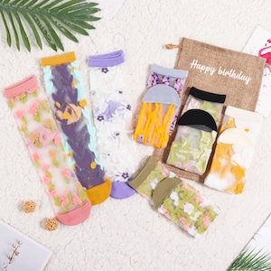 May include: A selection of sheer socks in various colours and floral designs. The socks feature pink, purple, yellow, and green accents. A burlap gift bag with "Happy birthday" printed on it is also visible.