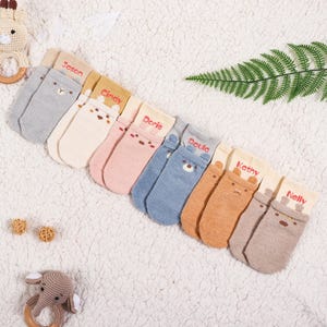Op de afbeelding: Acht paar babysokken met berengezichten en geborduurde namen. De sokken zijn in verschillende kleuren, waaronder grijs, beige, roze, blauw en bruin. De sokken zijn in een rij op een wit, pluche oppervlak gerangschikt.
