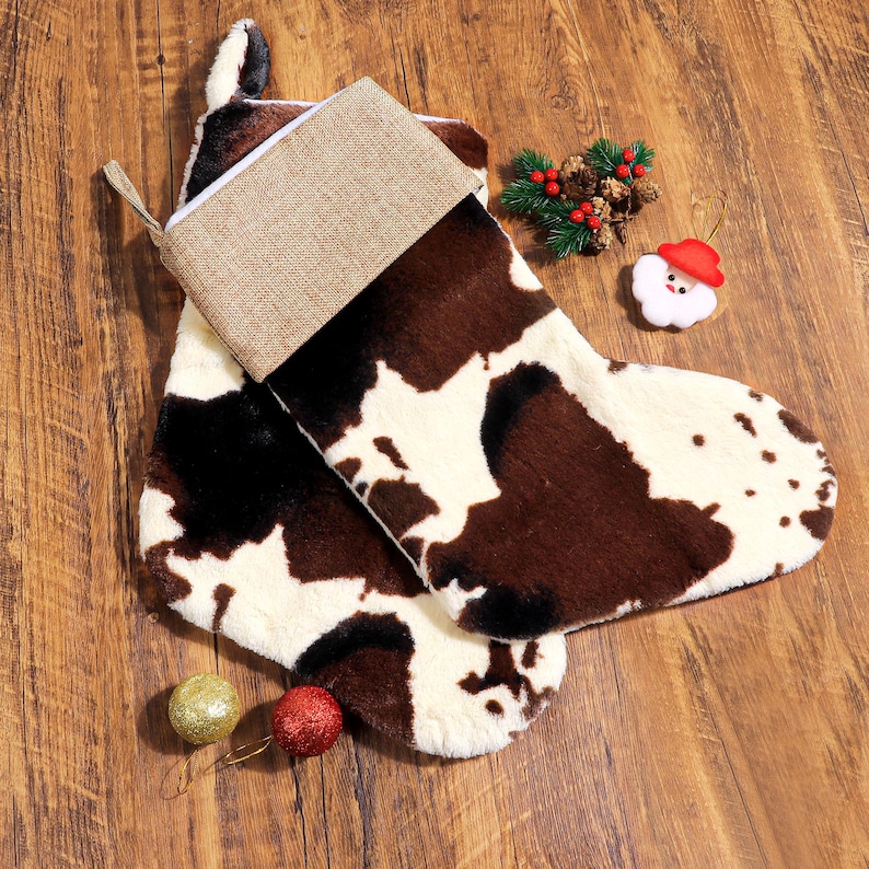 May include: A pair of Christmas stockings with a cowhide pattern in brown, black, and white. The top of the stocking is a tan burlap material. Decorative ornaments, including a Santa Claus and two glittery balls, are also present.