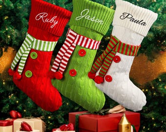 May include: Three Christmas stockings in red, green, and white, each with a name embroidered on the cuff. The stockings have cable knit patterns, striped scarves, and red buttons. They hang against a backdrop of Christmas tree branches.