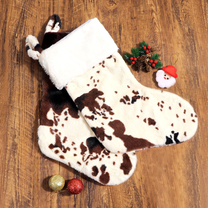 May include: Two Christmas stockings with a cowhide pattern in brown and white, trimmed with white faux fur. The stockings are on a wooden surface, with Christmas ornaments and a Santa Claus decoration.
