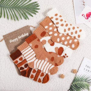 May include: A set of six cosy brown and white fuzzy socks with different patterns, including polka dots, stripes, and animal prints. The socks are arranged in a pile on a white surface with a hessian bag that says "Happy birthday" in white lettering.