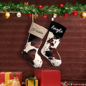 May include: Two Christmas stockings with a cowhide pattern, one with "Paula" embroidered on burlap and the other with "Angle" on black faux fur. The stockings hang from a garland with ornaments and stars.