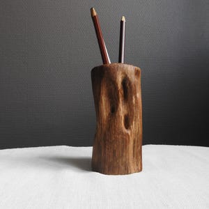 Peut inclure: Un porte-crayon cylindrique en bois fabriqué à partir d'une pièce de bois naturel avec une surface texturée et inégale et une finition brun foncé. Deux crayons brun foncé sont insérés dans l'ouverture supérieure du support.