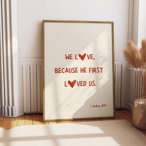 May include: A beige framed print with a white background and red text that reads "We love, because he first loved us. 1 John 4:19". The text includes red heart symbols.