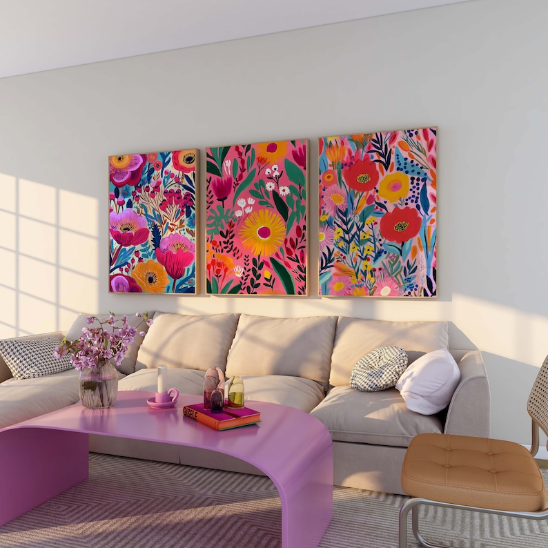 May include: Three colorful floral paintings with pink, red, yellow, and blue flowers on a white background hang above a beige sofa with pillows. A pink coffee table sits in front of the sofa.
