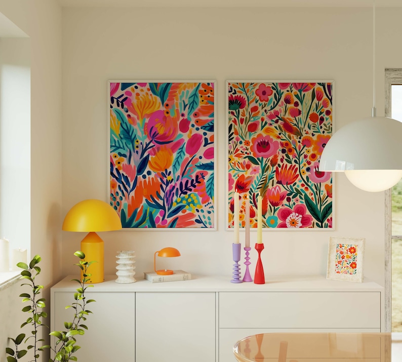 May include: Two framed floral prints with vibrant pink, orange, yellow, and green flowers on a white background. The prints are hanging on a white wall above a white dresser with a yellow lamp and candlesticks.