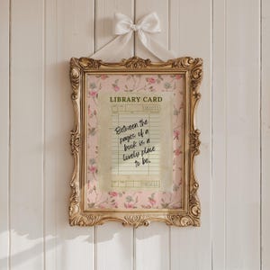 May include: Ornate gold framed print with a pink floral background. The print features a vintage library card design with the quote, "Between the pages of a book is a lovely place to be." A white bow hangs at the top.