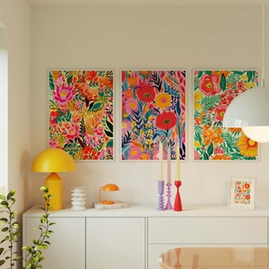 May include: Three framed floral prints with vibrant colors and patterns, hanging on a white wall above a white dresser with a yellow lamp and candlesticks.
