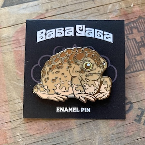 Puede incluir: Un pin de esmalte con una ilustración detallada de un sapo en tonos marrones y beige. El pin está sobre una tarjeta negra con las palabras "Baba Yaga" y "Enamel Pin".