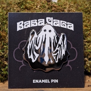 May include: A black enamel pin featuring a white ghost design with the text "Baba Yaga" above and "Enamel Pin" below. The pin is on a black card with a decorative border.