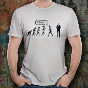 Puede incluir: Una camiseta blanca con una imagen en blanco y negro de una línea de figuras de palo que representan la evolución humana. La última figura de la línea es un hombre con traje, con un globo de diálogo sobre su cabeza que dice "Vuelve atrás, la hemos liado!"