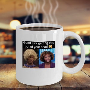 May include: White ceramic coffee mug with a black handle. The mug has a black rim and a printed image of two women with the text "Good luck getting this out of your head".