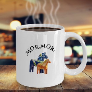 May include: White ceramic coffee mug with a black handle. The mug has a blue and orange Dala horse design and the text "MORMOR" in black.