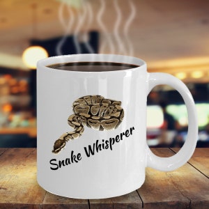 Könnte beinhalten: Weiße Keramiktasse mit einer Illustration einer zusammengerollten Schlange in Braun- und Beigetönen. Auf der Tasse steht in schwarzer Schrift der Text "Snake Whisperer". Die Tasse ist mit dunkler Flüssigkeit, möglicherweise Kaffee, gefüllt und steht auf einer Holzoberfläche.