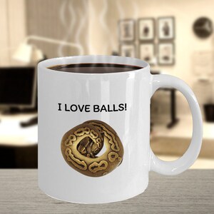 May include: White coffee mug with a brown snake curled up on it. The mug has the text "I LOVE BALLS!" printed on it.
