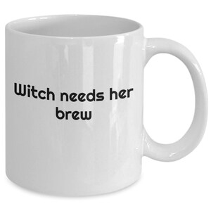Könnte beinhalten: Weißer Keramikbecher mit gebogenem Henkel. Der Becher trägt den schwarzen Text "Witch needs her brew" mittig auf der Vorderseite. Der Becher ist für Heißgetränke konzipiert.