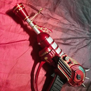 May include: A red and gold ray gun prop with a black handle and silver accents. The toy gun has a detailed design with a trigger, a barrel, and a scope-like component. The prop is likely for cosplay or display.