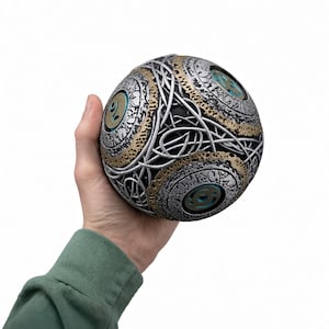 May include: A spherical object featuring a complex, textured surface with silver and gold metallic finishes. The design includes intricate, raised, interlaced lines and circular patterns with recessed teal-colored accents throughout the structure.