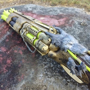 May include: A fantasy prop weapon with a gold and bronze finish. The design incorporates a dragon head and claw details, with neon green accents and tubes. A chain is attached to the weapon. The weapon is lying on a rock.