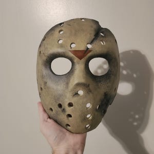 May include: A tan hockey mask with dark eye holes and numerous circular ventilation holes. The mask has a red triangle at the forehead and is weathered with dark smudges. A classic horror movie prop.