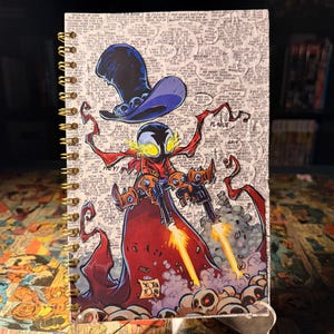 May include: Spiral-bound notebook with a comic book illustration. The cover features a character with a top hat, red cape, and guns firing. The background is filled with comic book text. Gold spiral binding.