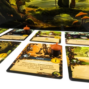 Puede incluir: Primer plano de un juego de mesa con cartas que muestran ilustraciones de una gr&uacute;a, un monasterio, una granja y una esposa. Las cartas est&aacute;n sobre una superficie blanca con fichas con forma de bellota.