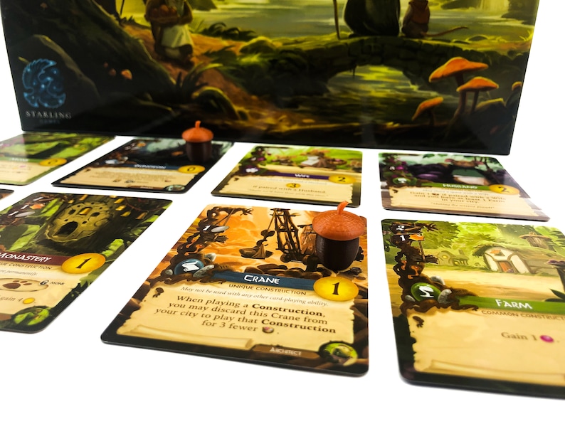 Puede incluir: Primer plano de un juego de mesa con cartas que muestran ilustraciones de una gr&uacute;a, un monasterio, una granja y una esposa. Las cartas est&aacute;n sobre una superficie blanca con fichas con forma de bellota.