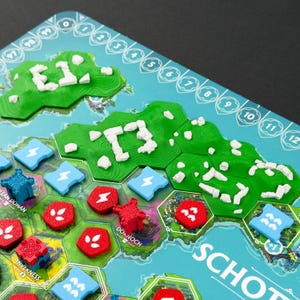 Könnte beinhalten: Ein Brettspiel mit dem Titel SCOTLAND, das eine Landkarte mit grünem Gelände, weißen Steinstrukturen und verschiedenen farbigen Spielsteinen in Rot, Blau und Hellblau zeigt. Das Spielbrett enthält eine nummerierte Leiste am Rand.