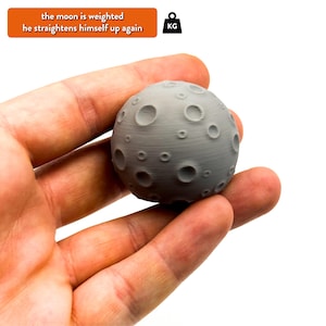 May include: A grey, 3D printed moon with craters, held in a hand. The moon is weighted and the text "the moon is weighted he straightens himself up again" is visible above the hand.