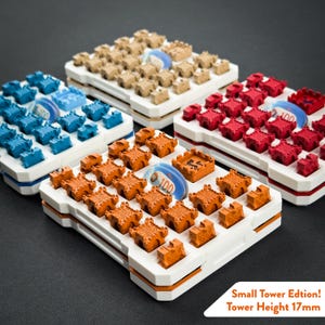 Könnte beinhalten: Vier rechteckige weiße Tabletts mit kleinen burgförmigen Spielfiguren in Beige, Blau, Rot und Orange. Jedes Tablett enthält einen blauen runden Spielstein mit der Aufschrift 100. Der Text lautet: Small Tower Edition! Tower Height 17mm.
