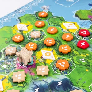 Puede incluir: Un juego de mesa con un mapa hexagonal verde y una pieza de castillo beige etiquetada como Dunguaire. El tablero incluye fichas naranjas y rojas con iconos blancos de rayos u hojas, losetas en forma de diamante y una pista numerada.