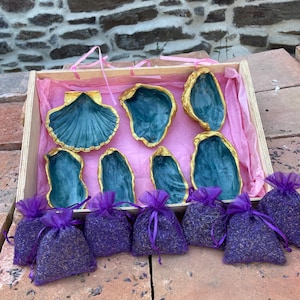 May include: A wooden gift box containing seven teal-colored seashell dishes with gold-painted edges, lined with pink tissue paper, and accompanied by six small purple organza drawstring bags filled with dried lavender buds.