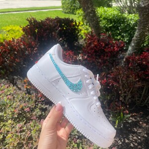 Puede incluir: Zapatillas bajas de cuero blanco con un swoosh de color azul turquesa adornado con joyas.