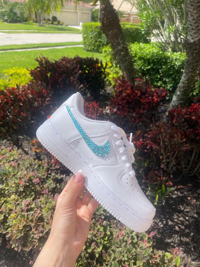 Puede incluir: Zapatillas bajas de cuero blanco con un swoosh de color azul turquesa adornado con joyas.