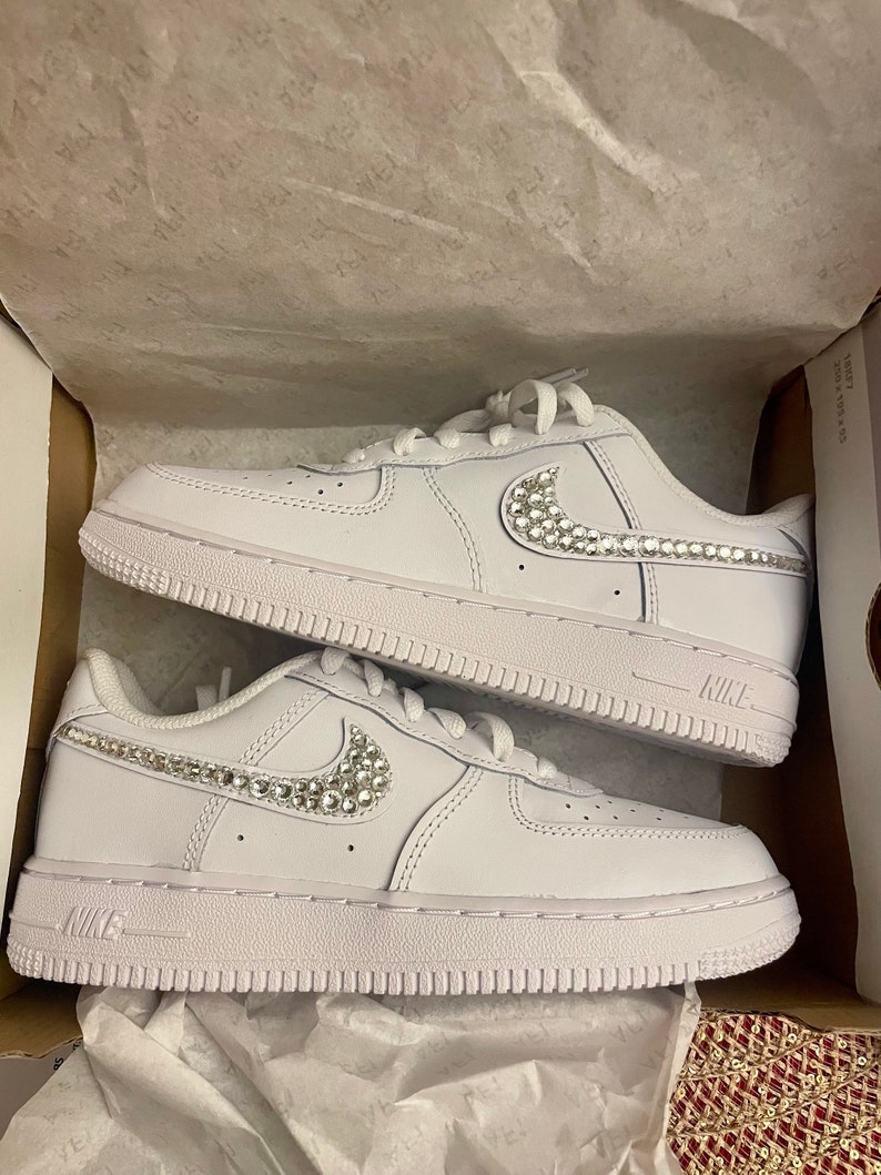 Puede incluir: Zapatillas bajas de cuero blanco con un logotipo swoosh blanco adornado con diamantes de imitaci&oacute;n. Las zapatillas est&aacute;n en una caja de zapatos blanca.